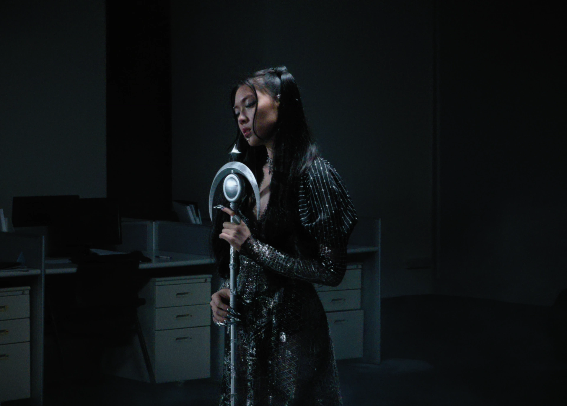 Jasmine Sokko in a metallic gown with a sculptural silver microphone in a dim room.