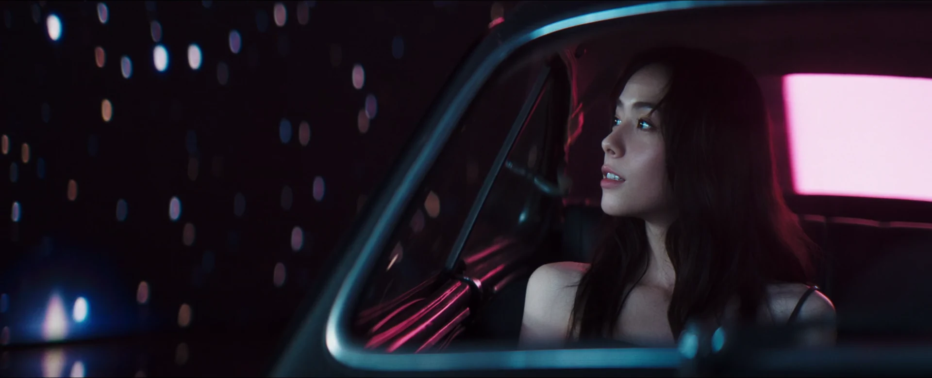 Charlize Lamb looks out of a vintage car window against pink and blue light.