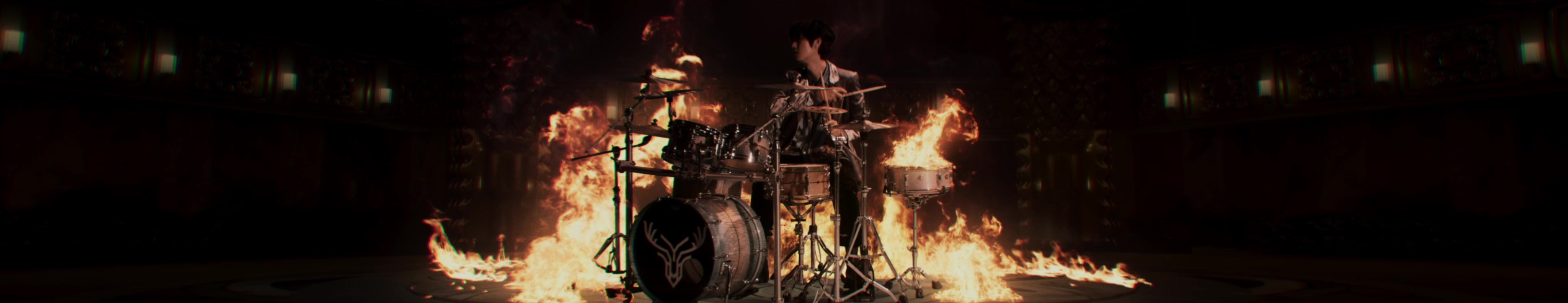 A drummer plays at a kit surrounded by rising flames.