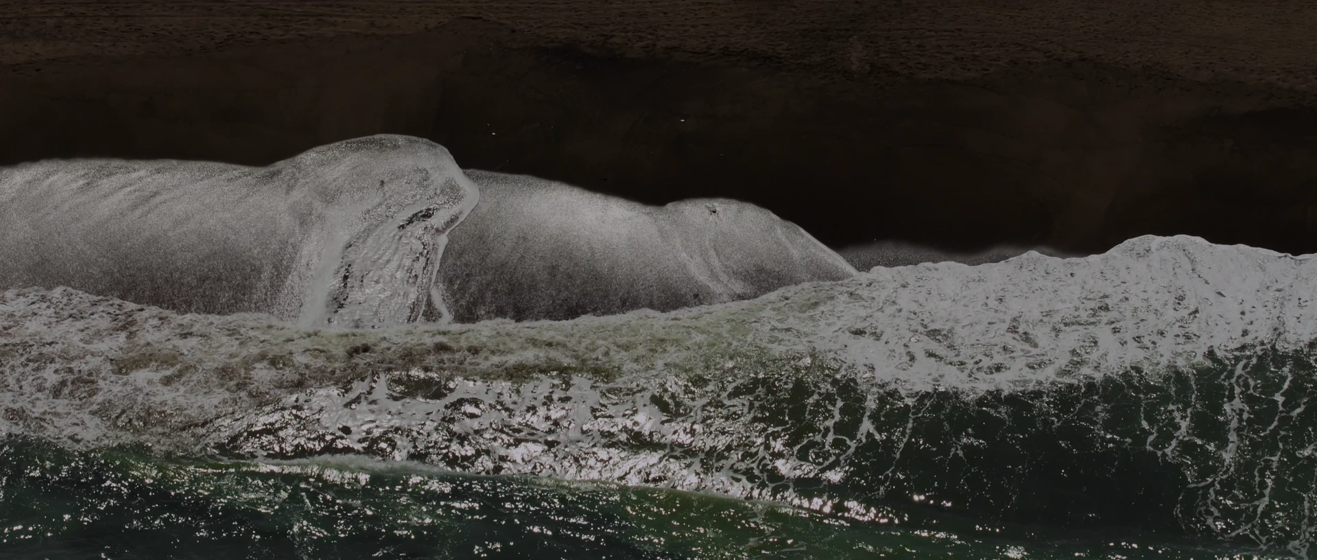 An aerial view of white surf folding onto dark sand.