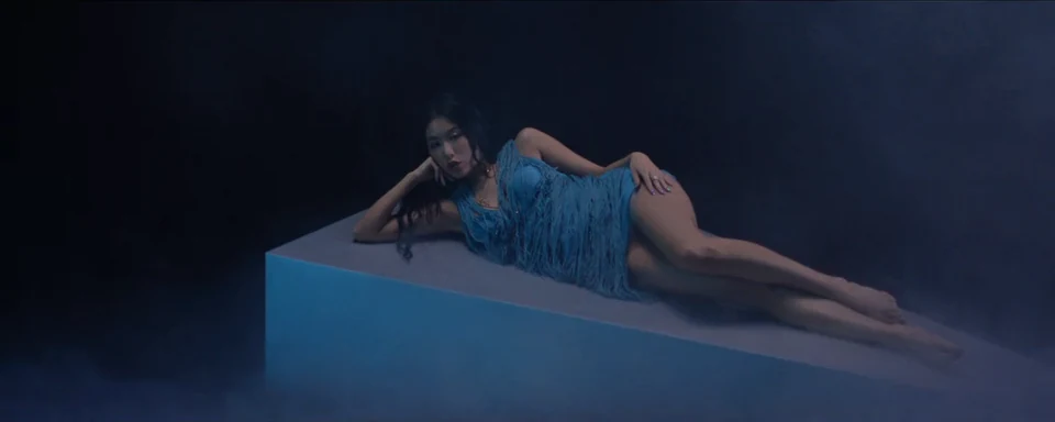Bella Su reclines on a pale blue plinth against a dark backdrop.